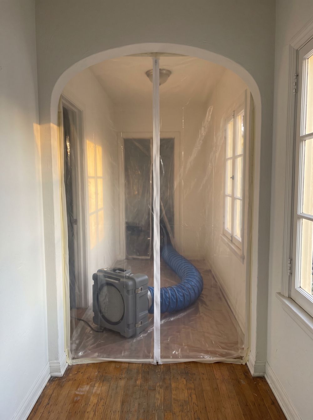 Clear plastic sheeting with a zippered door sealing off an arched hallway, an air scrubber and its duct behind it